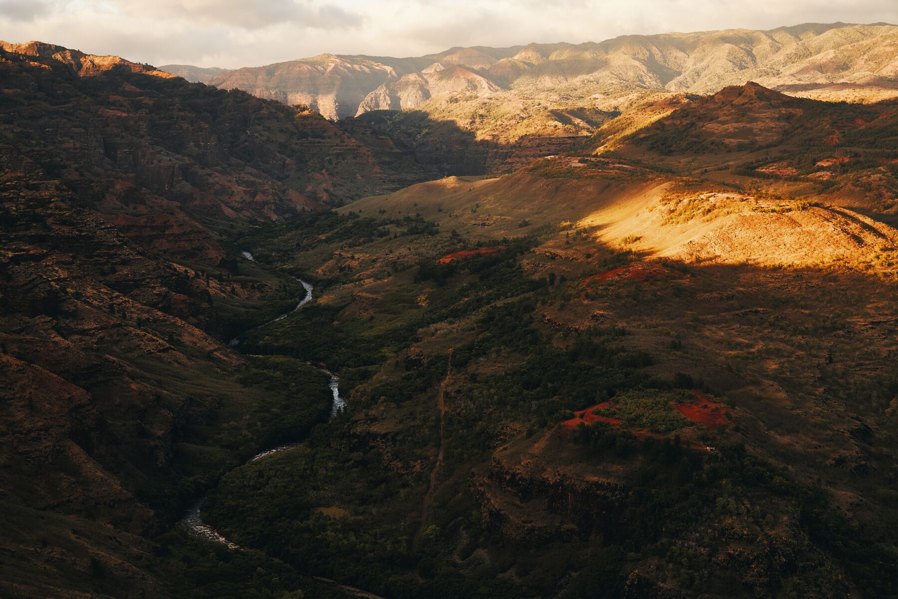 High-angle view into a deep, shadowed valley in Waimea Canyon, with a thin stream along the bottom and low sun lighting red-soil ridges on the right