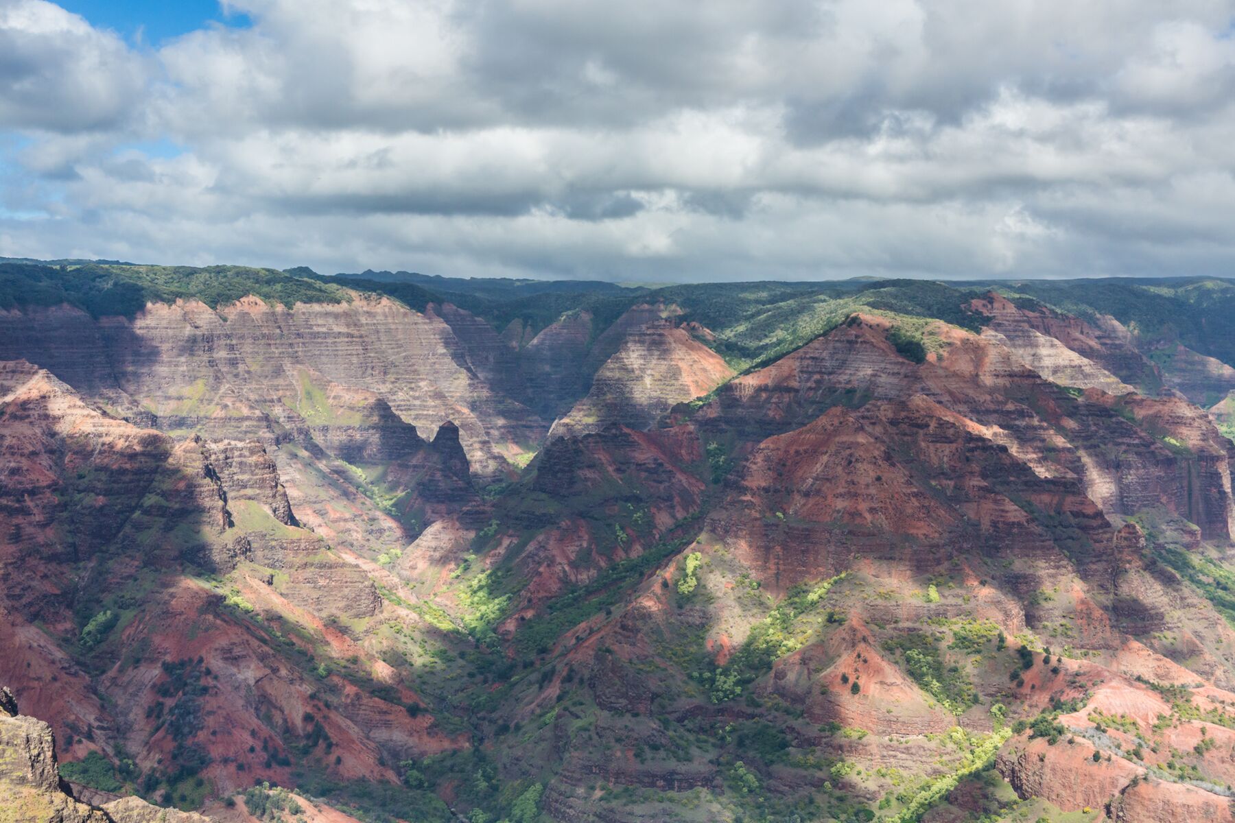 Layered red, purple-brown and green ridges fill Waimea Canyon from side to side under a sky of heavy white clouds