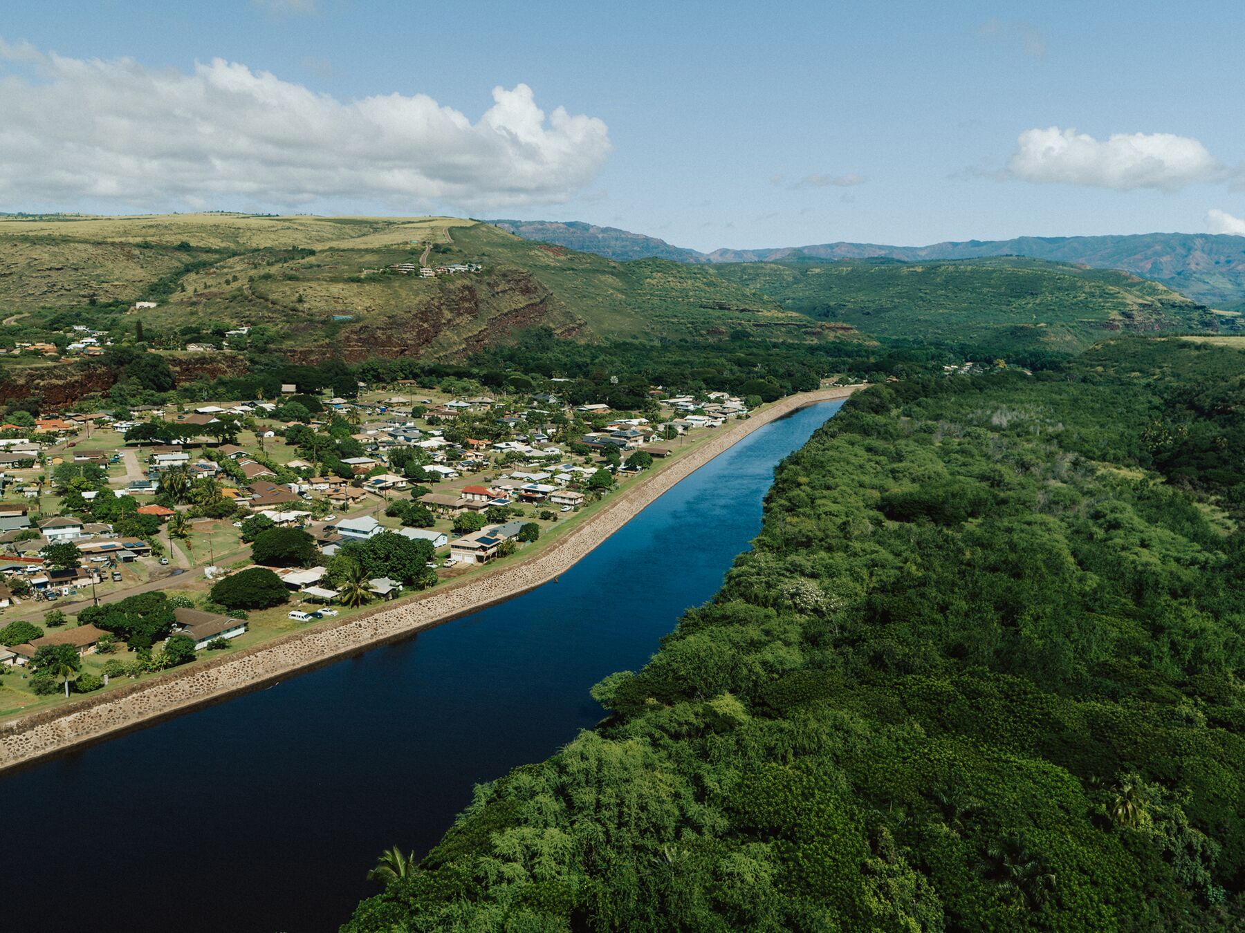 Aerial view of Waimea: houses and trees on one bank of a wide, dark-blue river, forest on the other, and green hills beyond