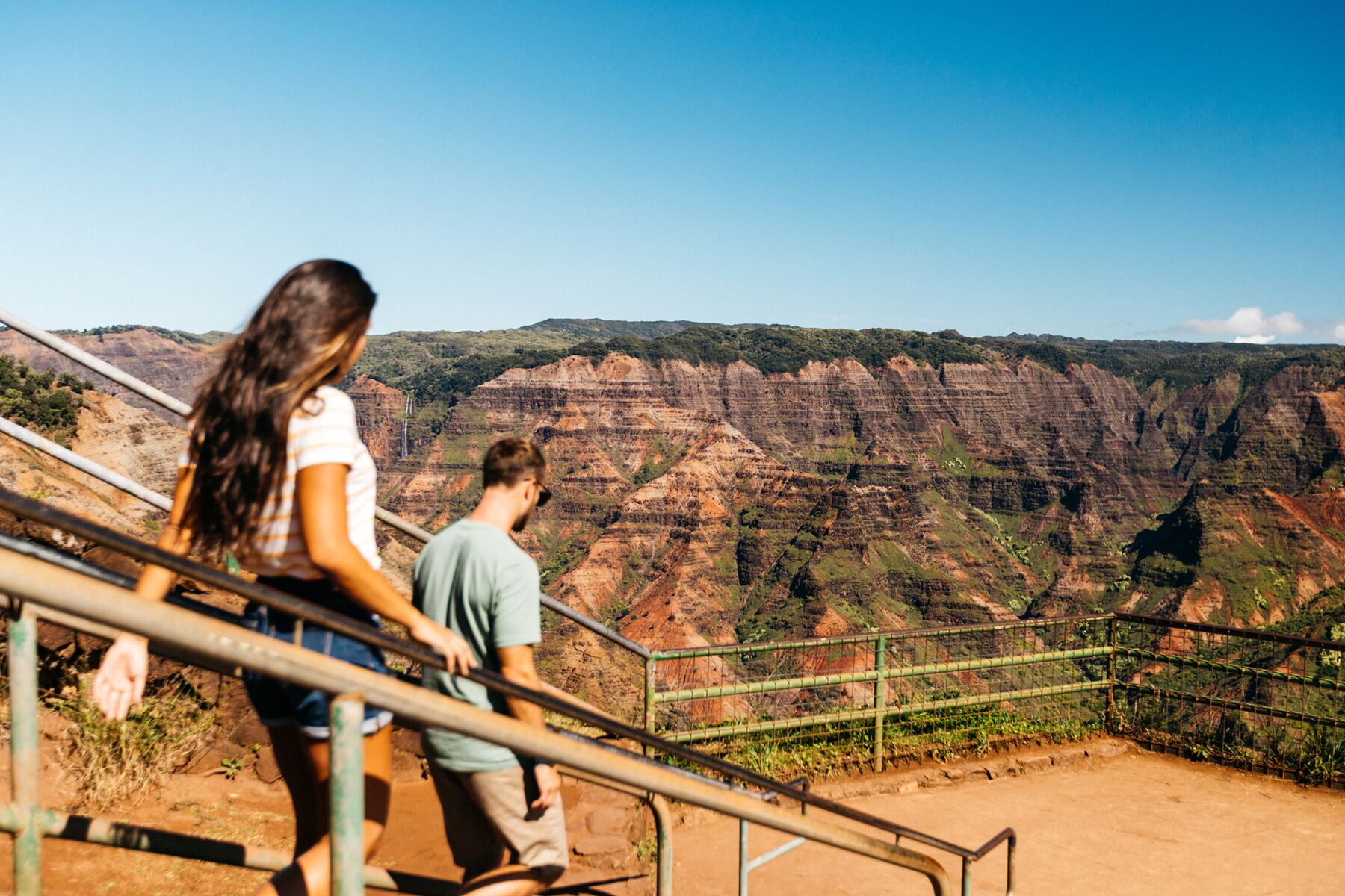 A woman and a man walk down steps with handrails toward the Waimea Canyon Lookout platform, with the canyon’s layered walls ahead