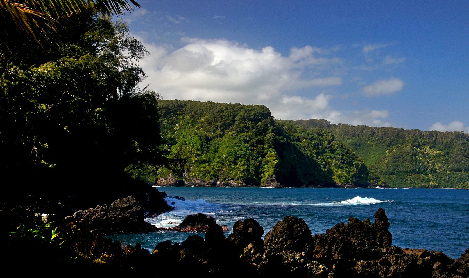 The Keʻanae Peninsula, a low point of black lava with surf breaking, under steep green cliffs