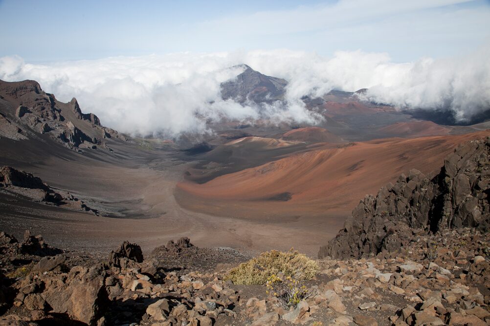 Clouds spilling over the rim into Haleakalā crater, above red cinder slopes and dark lava rock