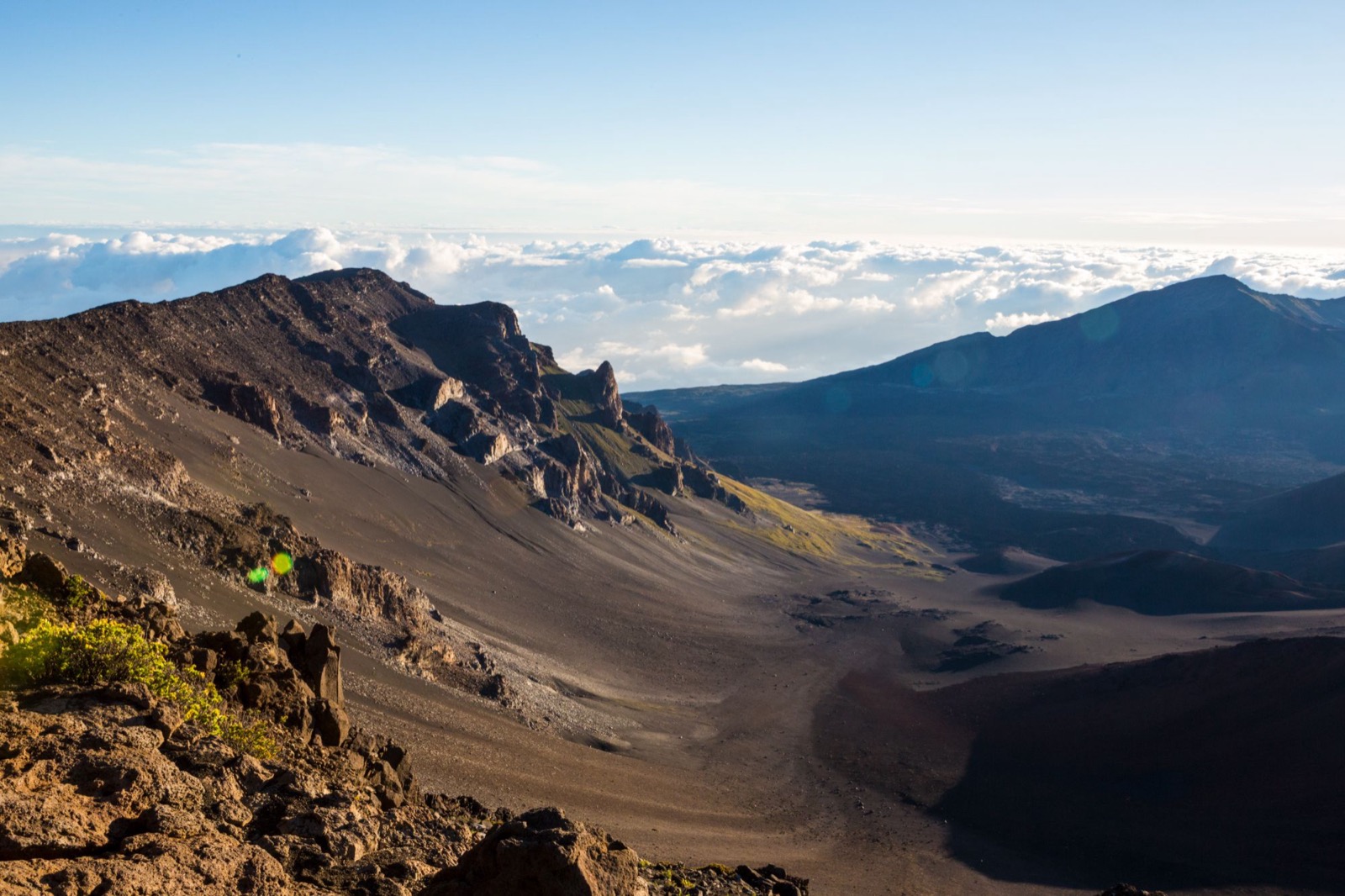 Morning light across Haleakalā crater’s rocky rim, cinder slopes and dark cinder cones, with a sea of clouds beyond