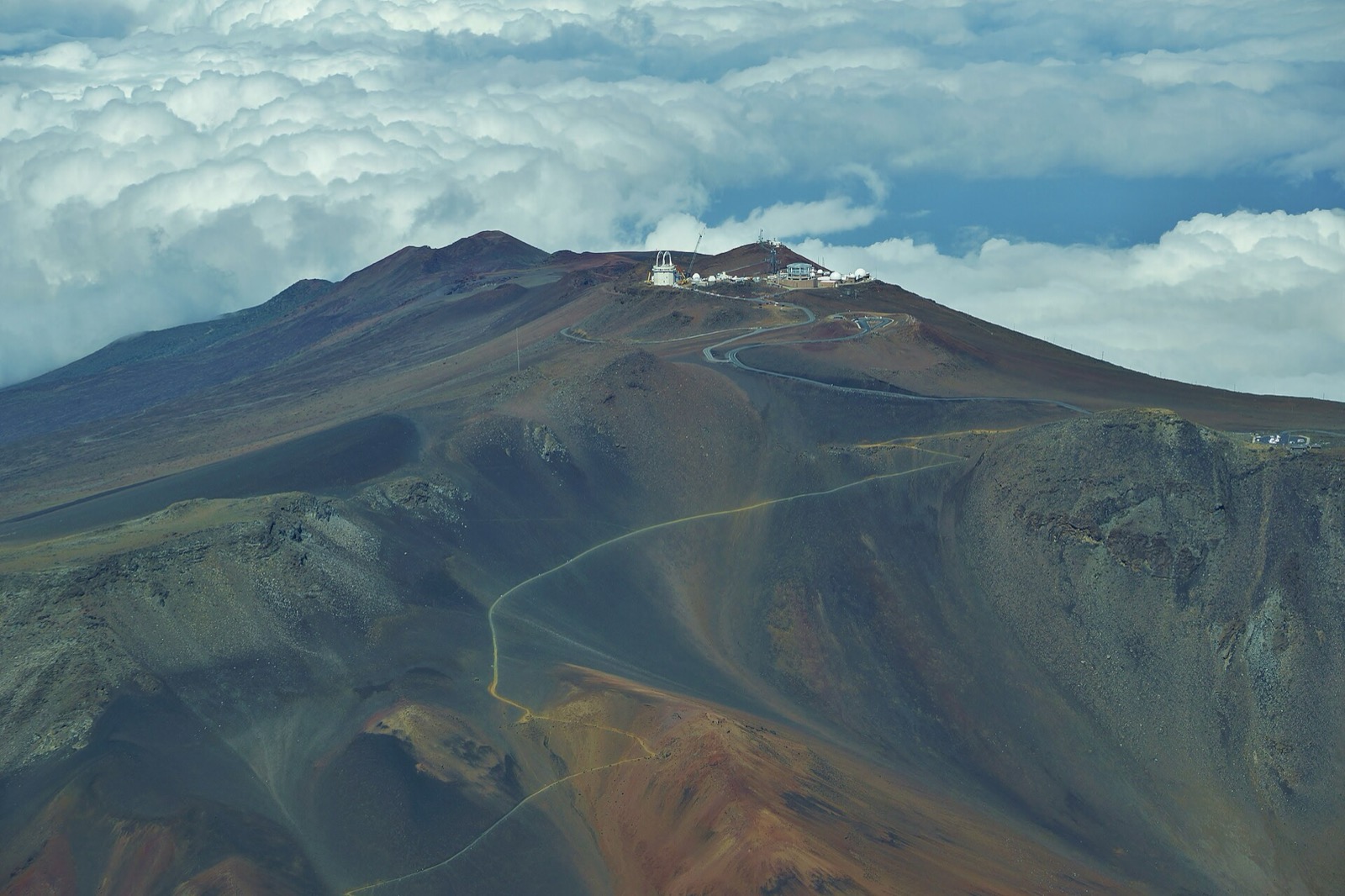 A trail winding across rust, charcoal and green cinder slopes toward the observatory domes on Haleakalā’s summit, above a sea of clouds