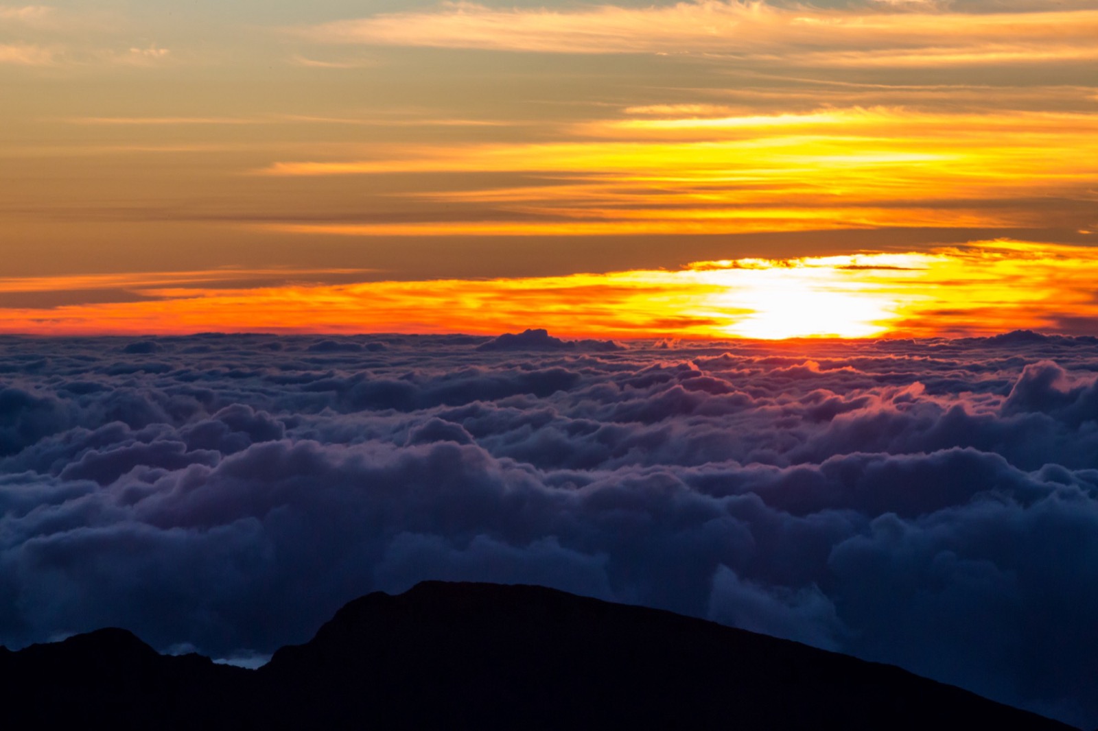 The sun breaking through bands of orange cloud above a sea of clouds, with a dark ridge silhouetted in the foreground