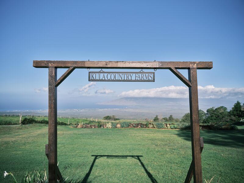 Wooden gateway with a Kula Country Farms sign framing green lawns and the view across Maui’s central valley to the West Maui Mountains