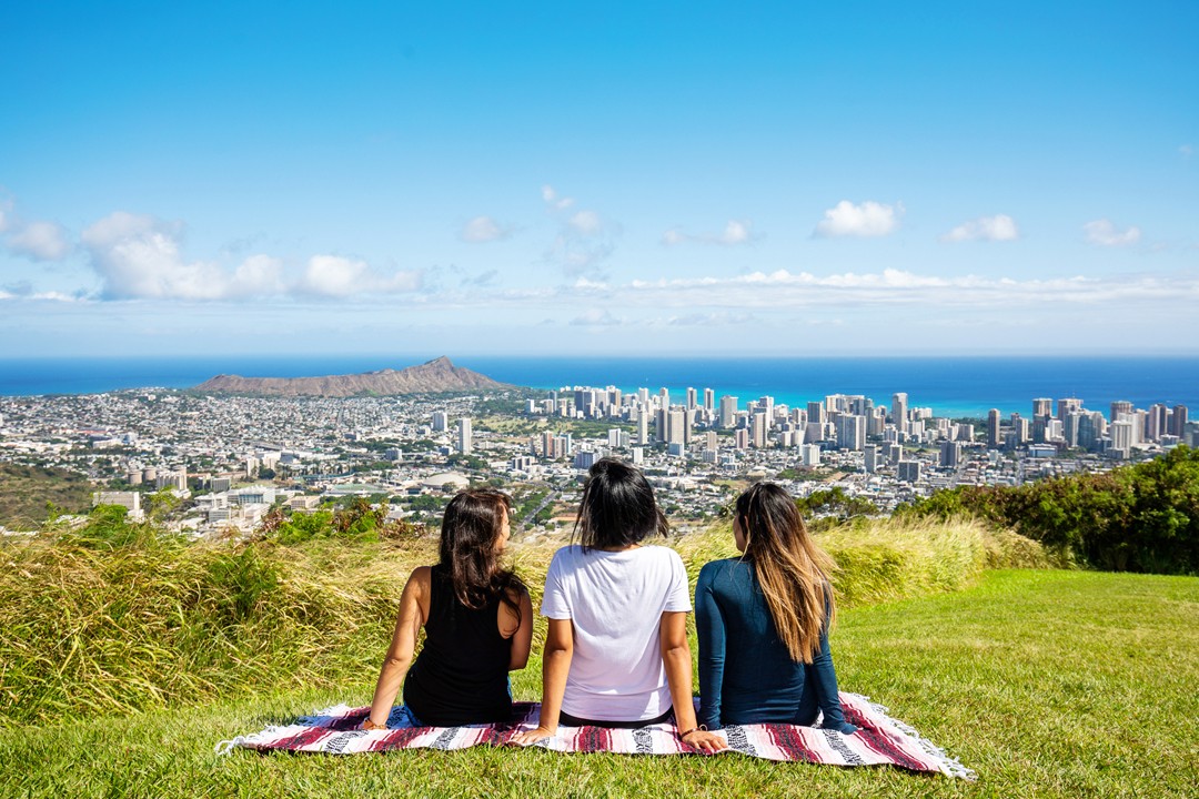 Three people on a blanket on a grassy hilltop looking out over Honolulu, Waikīkī and Lēʻahi